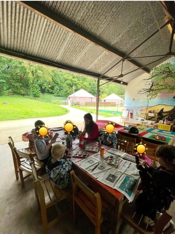 Atelier peinture sous le préau : les enfants, visages masqués, peignent autour d'une grande table, les yourtes en arrière-plan