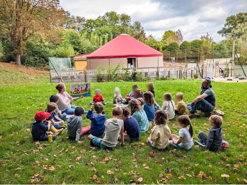 Lecture d'un album aux enfants assis en cercle sur la pelouse, devant une yourte